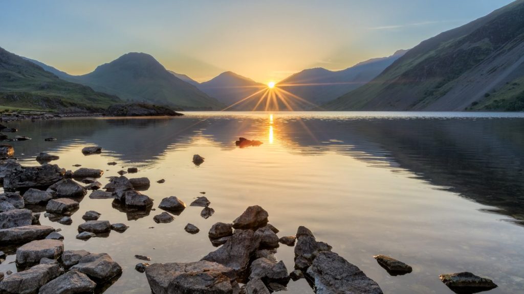 A photograph taken at Wastwater in the English Lake District on a fresh summer morning. The sun can be seen rising through the peaks of the mountains, creating a starburst sun ray effect.
